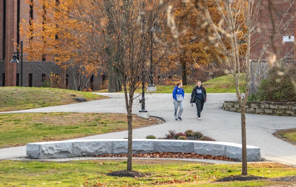 Two U N E students walk on a paved path through the greenery of Biddeford Campus